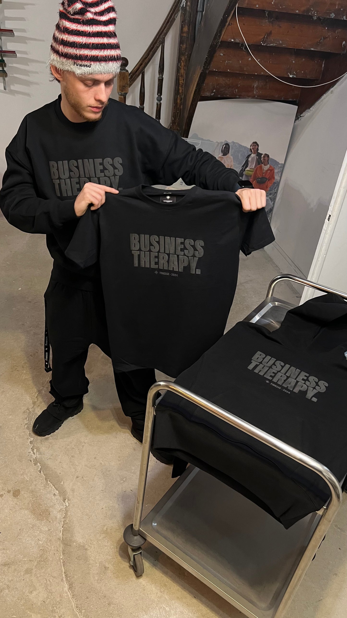 Homme tenant un T-shirt Black on Black, hoodie assorti posé sur chariot métallique.