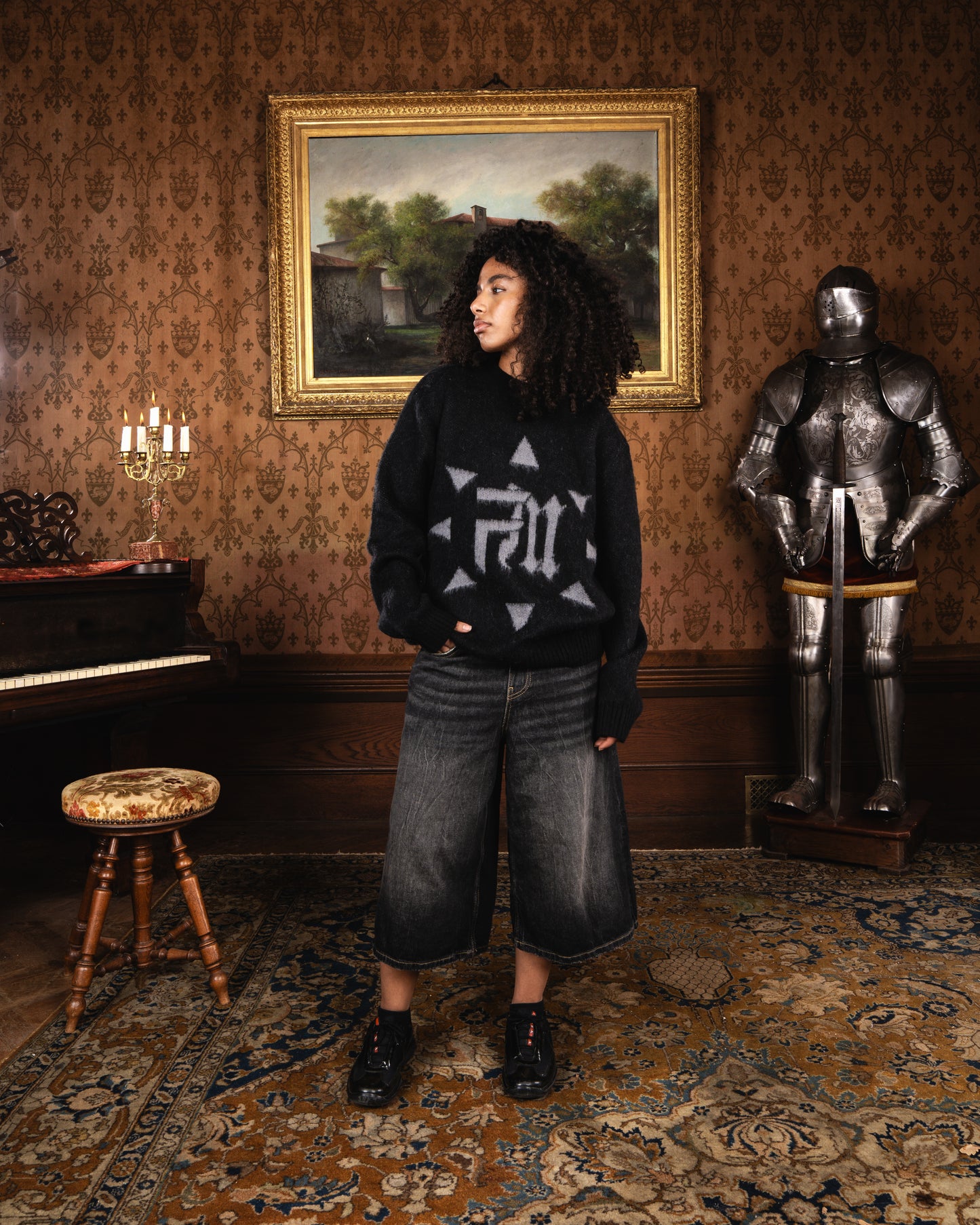 Femme en Finesse Mohair Black Bird posant devant un chandelier allumé et tapisserie orange, regard tourné de côté.