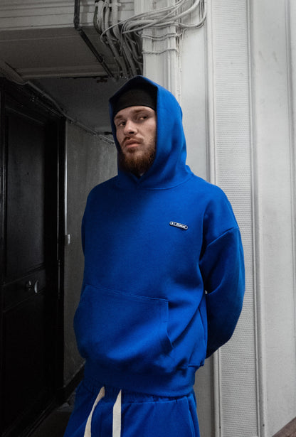 Homme barbu en bonnet portant le Finesse Air Cotton Hoodie bleu, regard pensif dans un bâtiment.