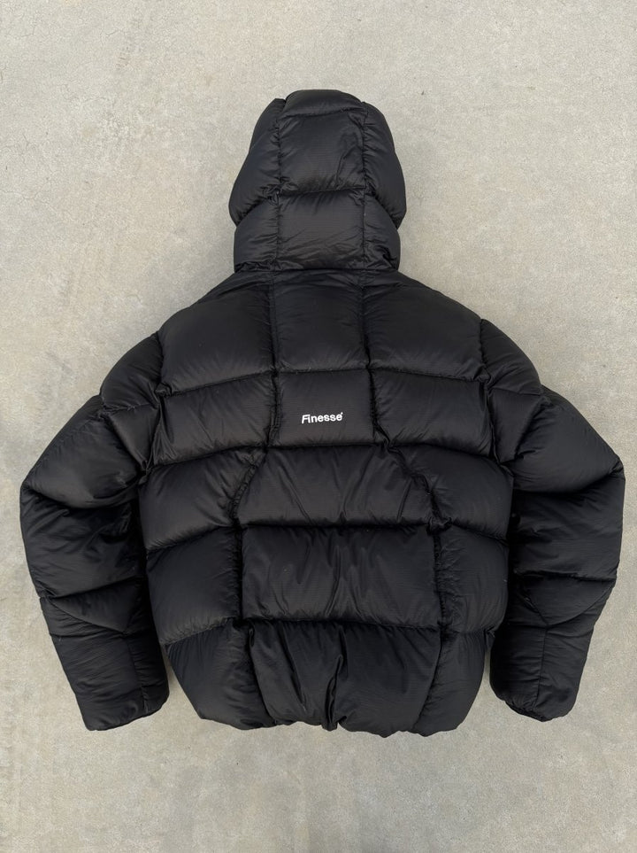 Alive Puffer Jacket noire posée au sol côté dos, sans chaussures visibles