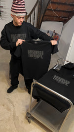 Homme tenant un T-shirt Black on Black, hoodie assorti posé sur chariot métallique.