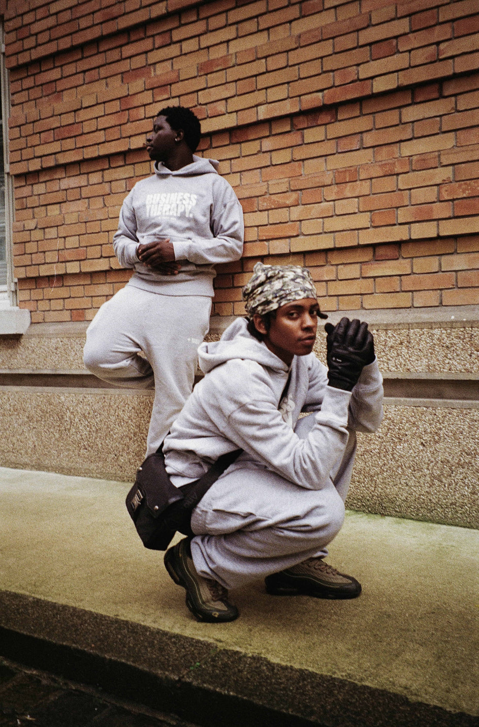 Deux hommes en jogging gris Business Therapy, l’un accroupi mains jointes, l’autre adossé au mur de briques
