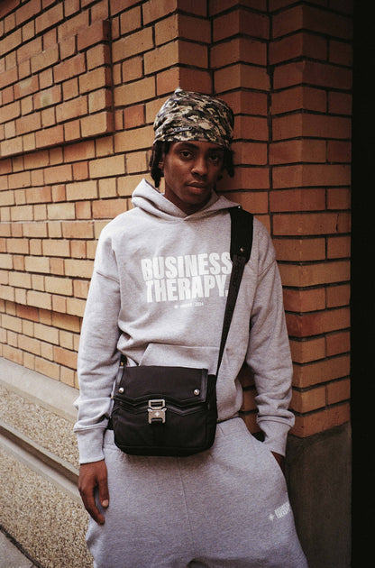 Homme cagoulé en jogging gris Business Therapy, au coin de la rue devant mur de briques