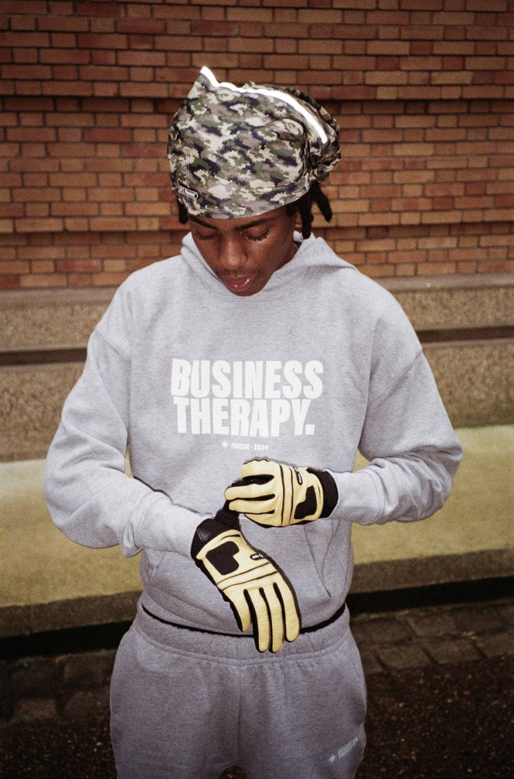 Homme en jogging gris Business Therapy, cagoule et Leather Gloves, posant devant mur de briques