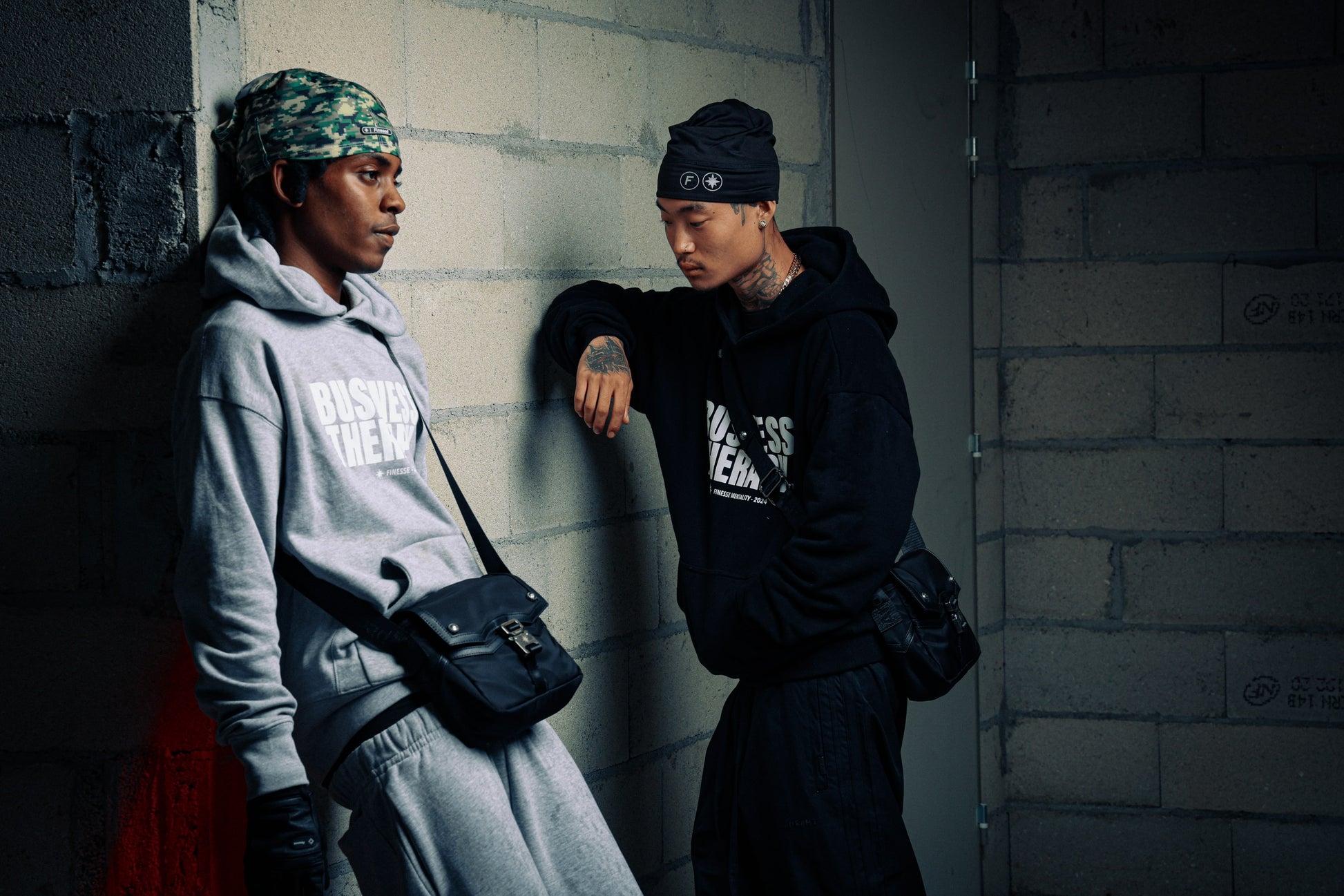 Deux hommes adossés à un mur en jogging noir Business Therapy, tous deux avec Memorial Bag.