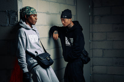 Deux hommes adossés à un mur en jogging noir Business Therapy, tous deux avec Memorial Bag.