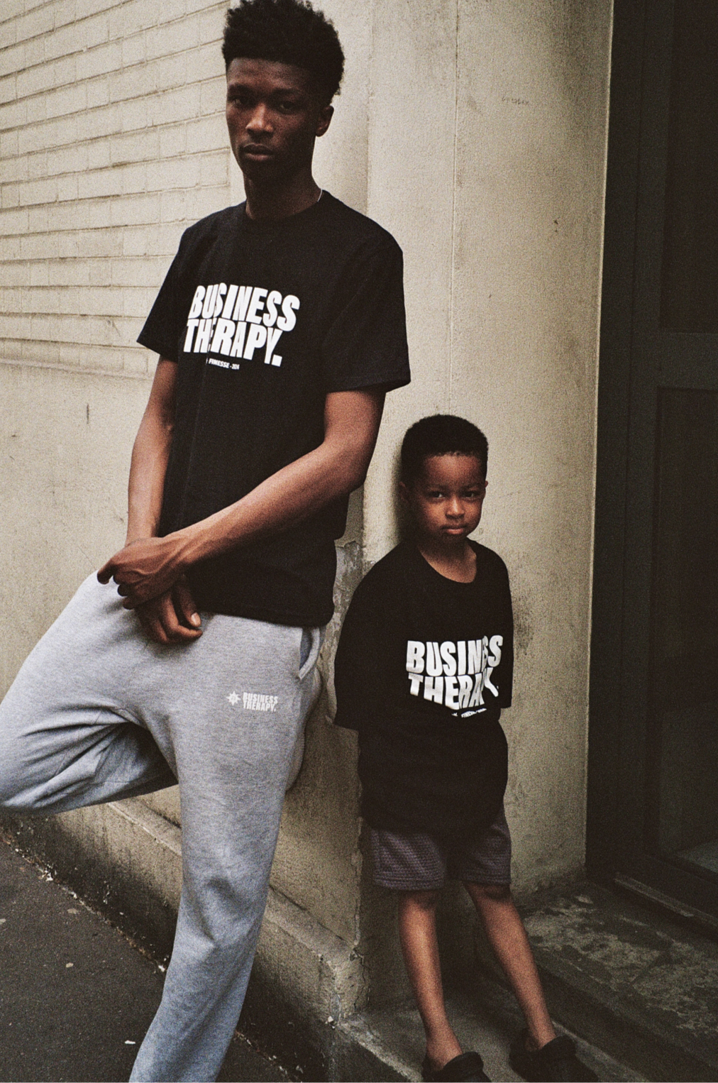 Homme et enfant adossés à un mur, portant tous deux un T-shirt Business Therapy.