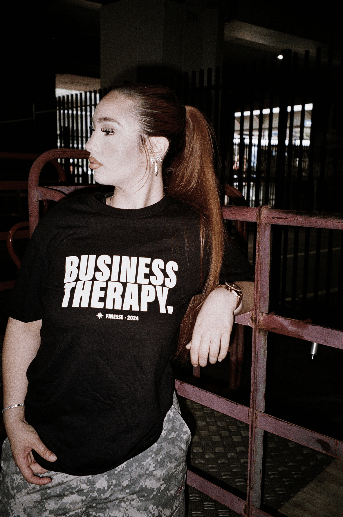 Femme dans un décor urbain portant un t-shirt noir Business Therapy, regard tourné vers la droite.