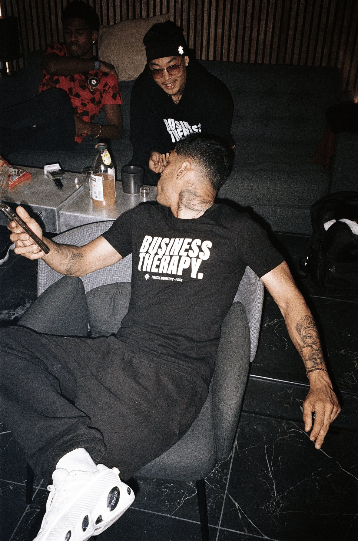 Homme tatoué allongé dans un canapé, portant un t-shirt Business Therapy et des sneakers blanches.