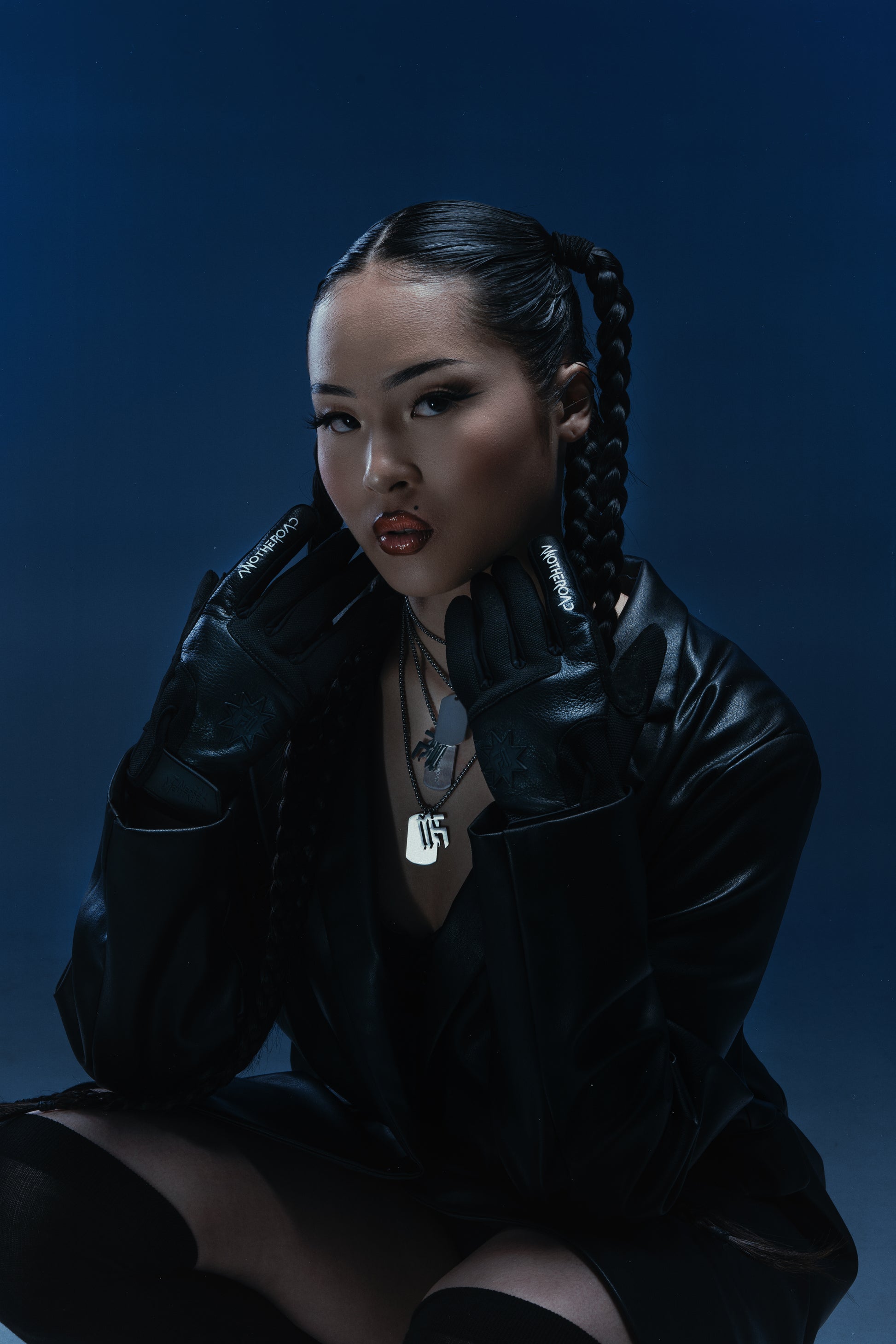 Femme assise aux tresses en veste cuir noir, portant la chaîne Finesse Qui Chaine et des gants Finesse Leather Gloves, sur fond bleu.