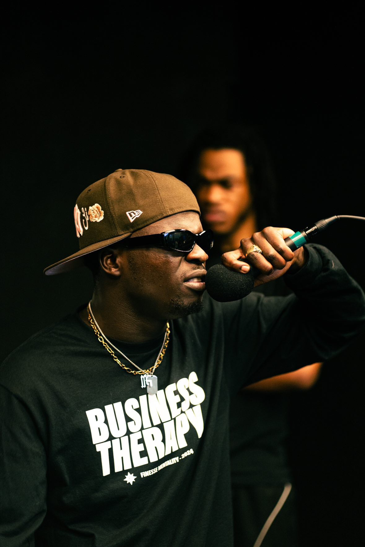 Homme sur scène avec casquette marron retournée, micro en main gauche, portant la chaîne Finesse et un long-sleeve Business Therapy.