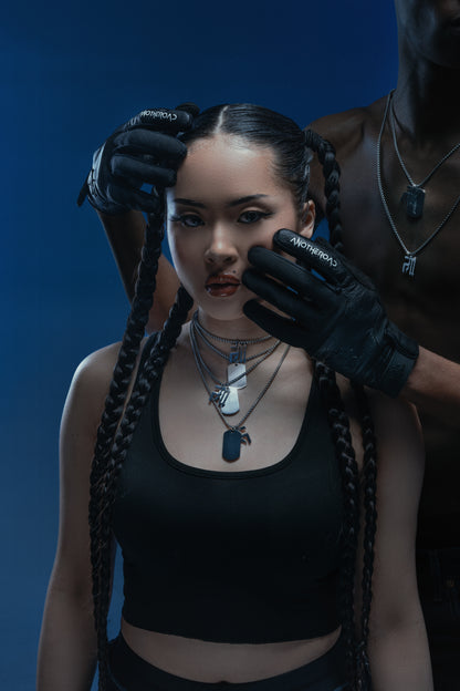 Femme en crop top noir portant quatre chaînes Finesse, coiffée de tresses, touchée au visage par un homme torse nu aux gants Finesse et chaînes.