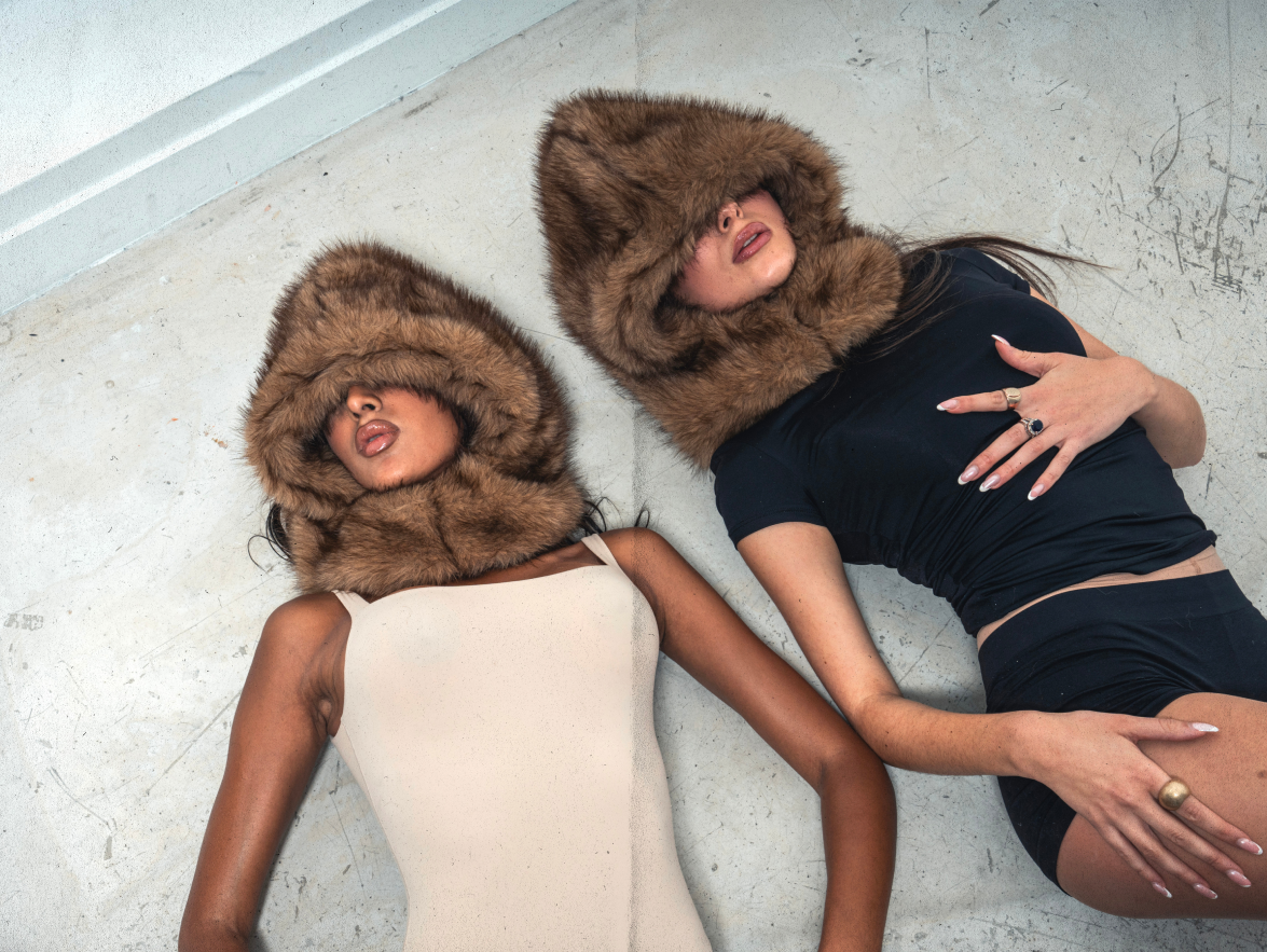 Deux femmes allongées au sol, l’une en beige, l’autre en noir, toutes deux avec FUR HOOD couvrant le visage.