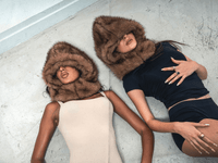 Deux femmes allongées au sol, l’une en beige, l’autre en noir, toutes deux avec FUR HOOD couvrant le visage.