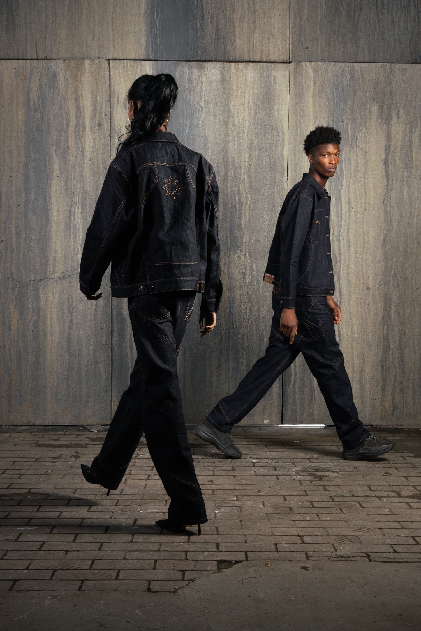 Un homme et une femme marchant dans un studio au mur industriel, tous deux en Finesse Denim Set ADV.