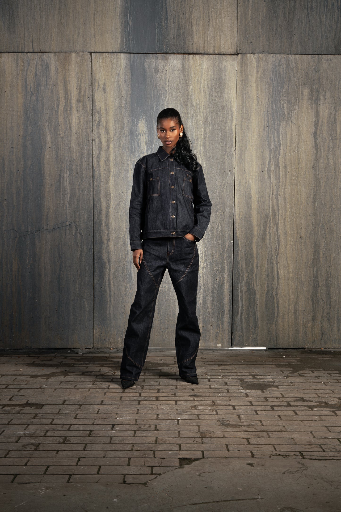 Femme de face en studio décor industriel, main dans la poche, portant le Finesse Denim Set ADV avec allure affirmée.