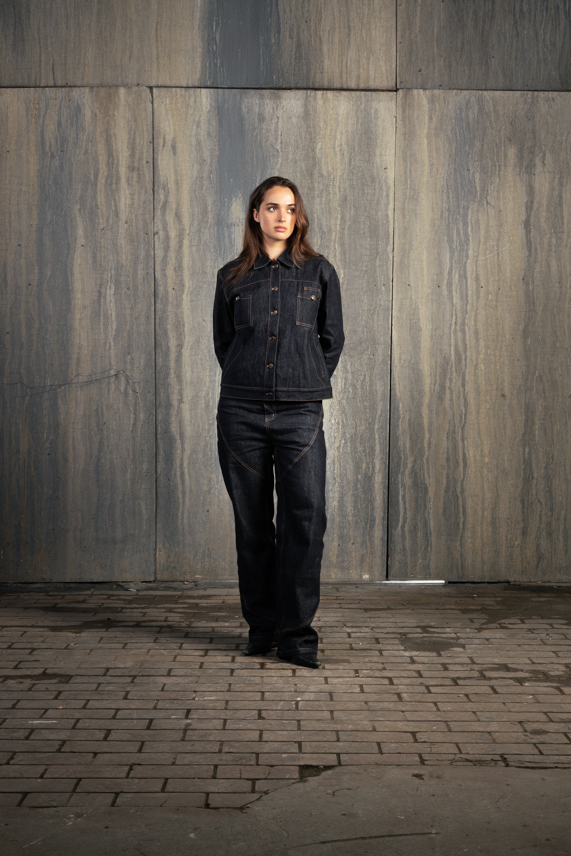 Femme en studio décor industriel, regard malicieux vers la gauche, mains derrière le dos, portant l’ensemble Finesse Denim Set ADV.