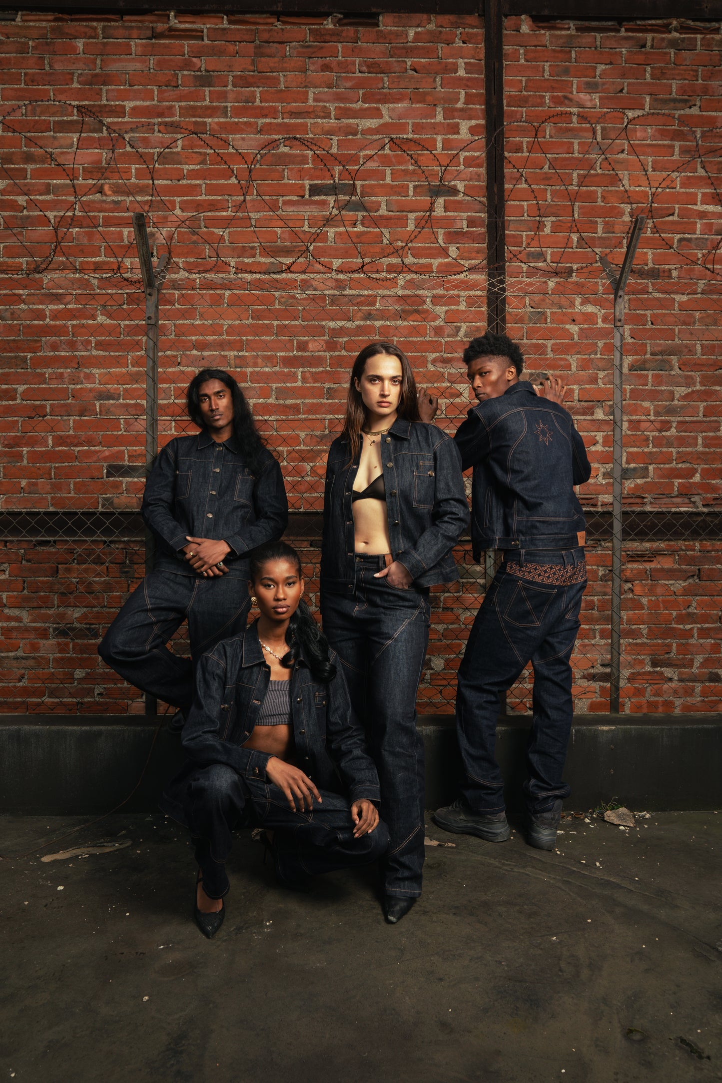Quatre mannequins devant une grille à barbelés et un mur de briques rouges, posant en Finesse Denim Set ADV.