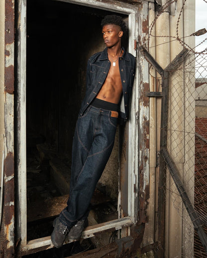 Homme torse nu en Finesse Denim Set veste ouverte, portant la Finesse Keychain, adossé à un encadrement de porte abîmé.