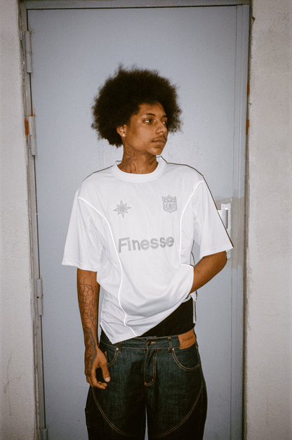 Homme tatoué devant une porte portant un Finest Denim Short et un Finesse Jersey Shirt blanc.