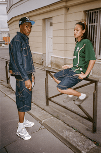 Femme assise sur une barrière en Finest Denim Short et Jersey vert, avec un homme en Denim Short, veste et casquette.