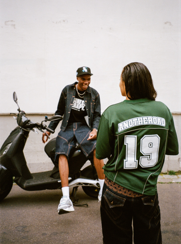 Plan large avec une femme en Jersey Shirt vert Finesse et un homme riant sur un scooter en Denim Vest Finesse.
