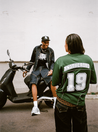 Plan large avec une femme en Jersey Shirt vert Finesse et un homme riant sur un scooter en Denim Vest Finesse.