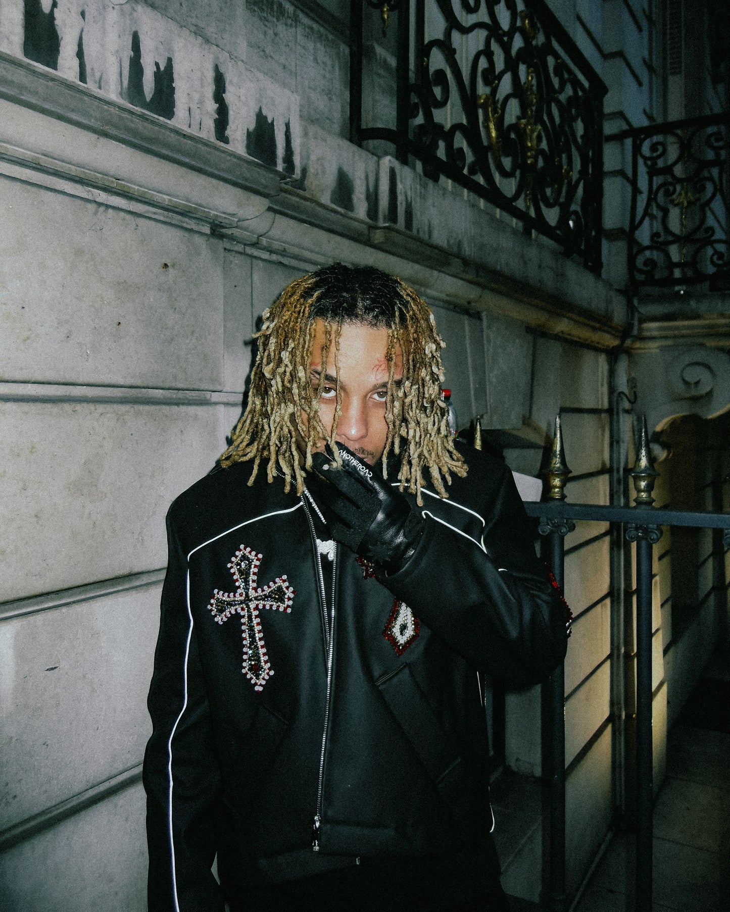 Homme aux dreadlocks blondes en veste cuir à croix, portant les gants Finesse Leather Gloves devant un immeuble haussmannien.
