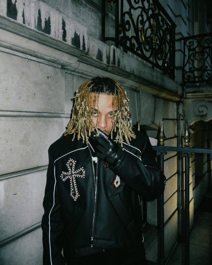 Homme aux dreadlocks blondes en veste cuir à croix, portant les gants Finesse Leather Gloves devant un immeuble haussmannien.