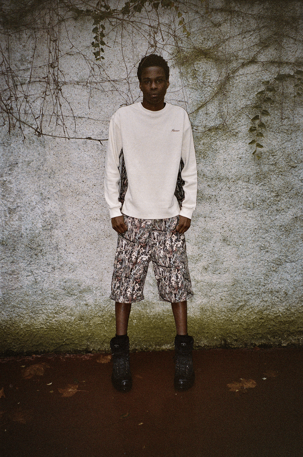 Homme adossé à un mur blanc-vert, portant le Finesse Military Top-Camo et le 2-in-1 Cargo Pants-Camo en version short.