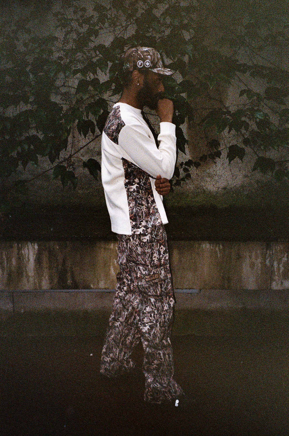 Homme de profil avec main sur le menton, en Finesse Military Top-Camo, 2-in-1 Cargo Pants-Camo et "SIGN" Cap-CAMO.