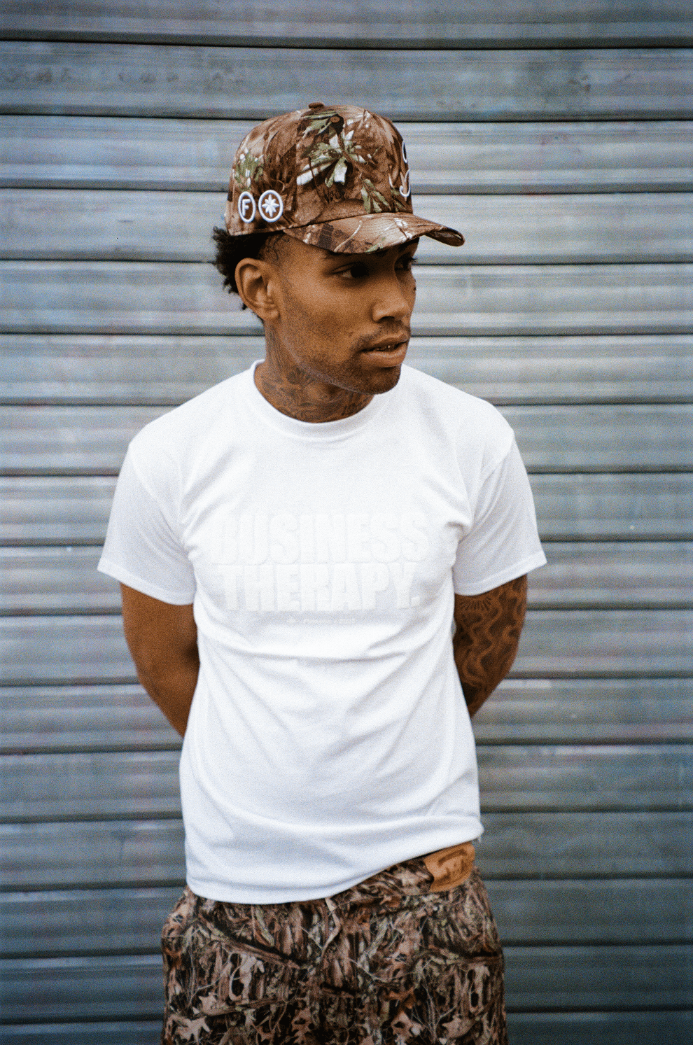 Homme bras derrière le dos, portant la Finesse "SIGN" Cap-CAMO et le Business Therapy T-shirt Puff White, fond rideau de fer.