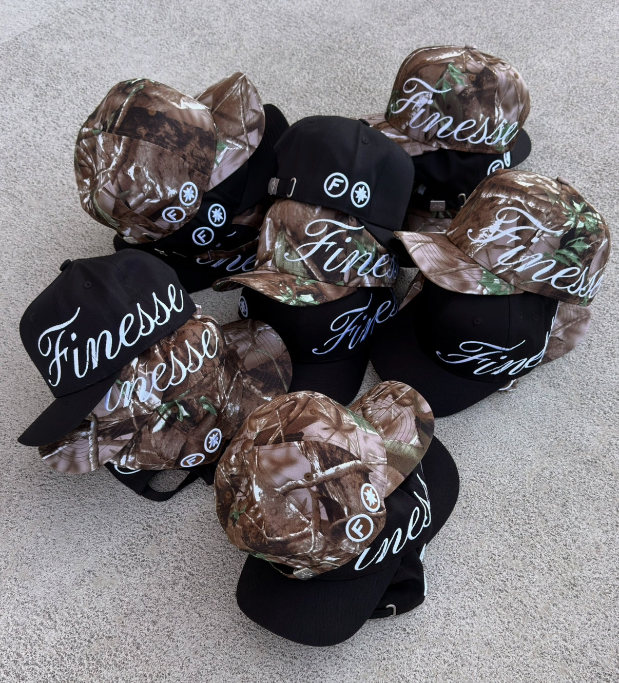 Packshot de plusieurs Finesse "SIGN" Cap-CAMO et "SIGN" Cap-Black alignées au sol, casquettes streetwear signature Finesse.