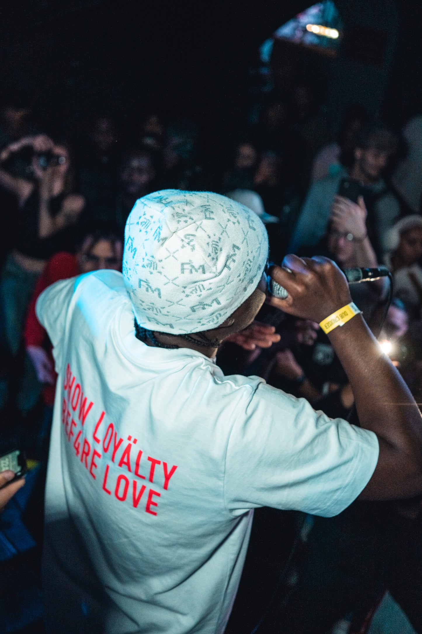 Homme de dos sur scène en t-shirt blanc et bonnet Finesse Snow, micro à la main durant un concert.