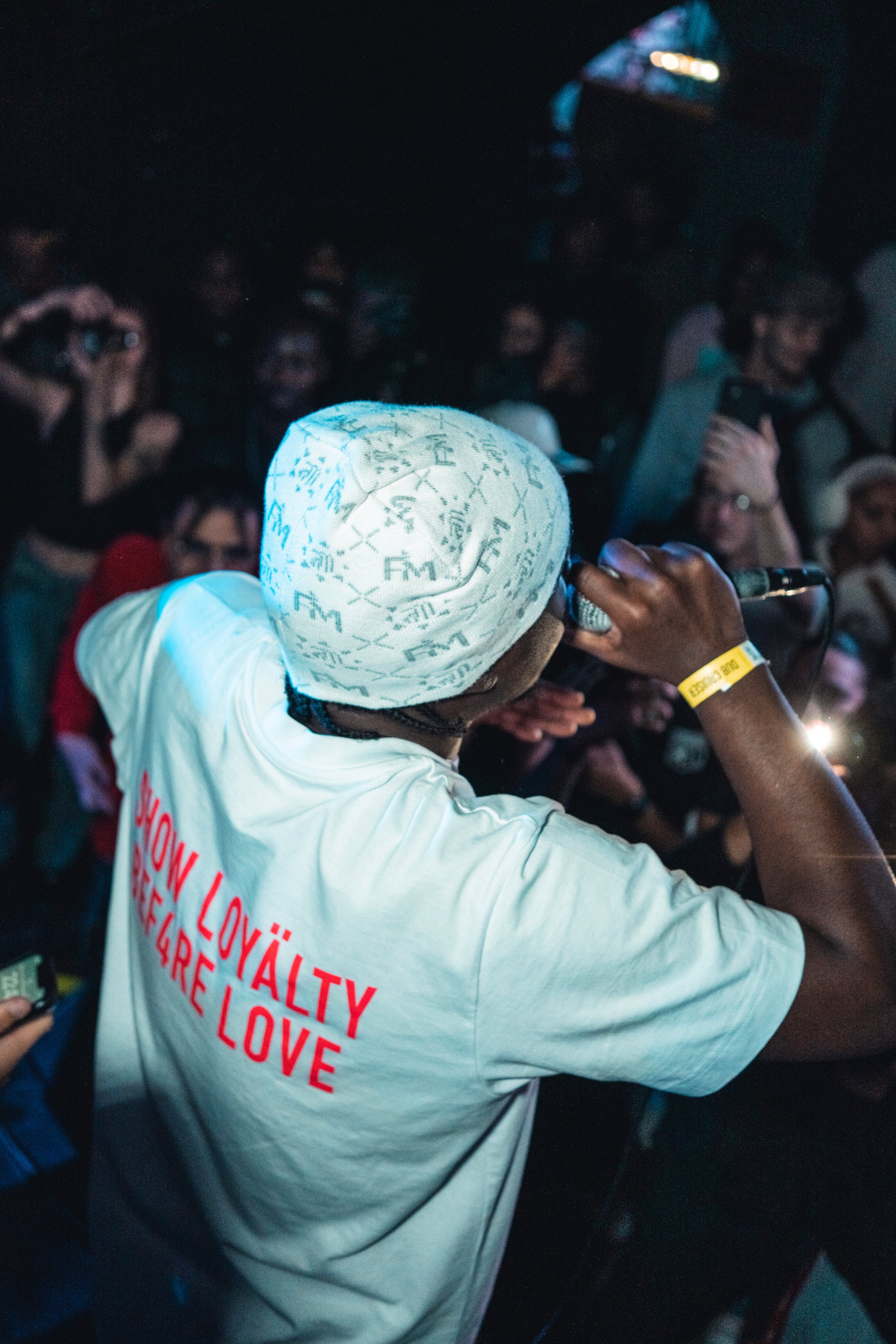 Homme de dos sur scène en t-shirt blanc et bonnet Finesse Snow, micro à la main durant un concert.