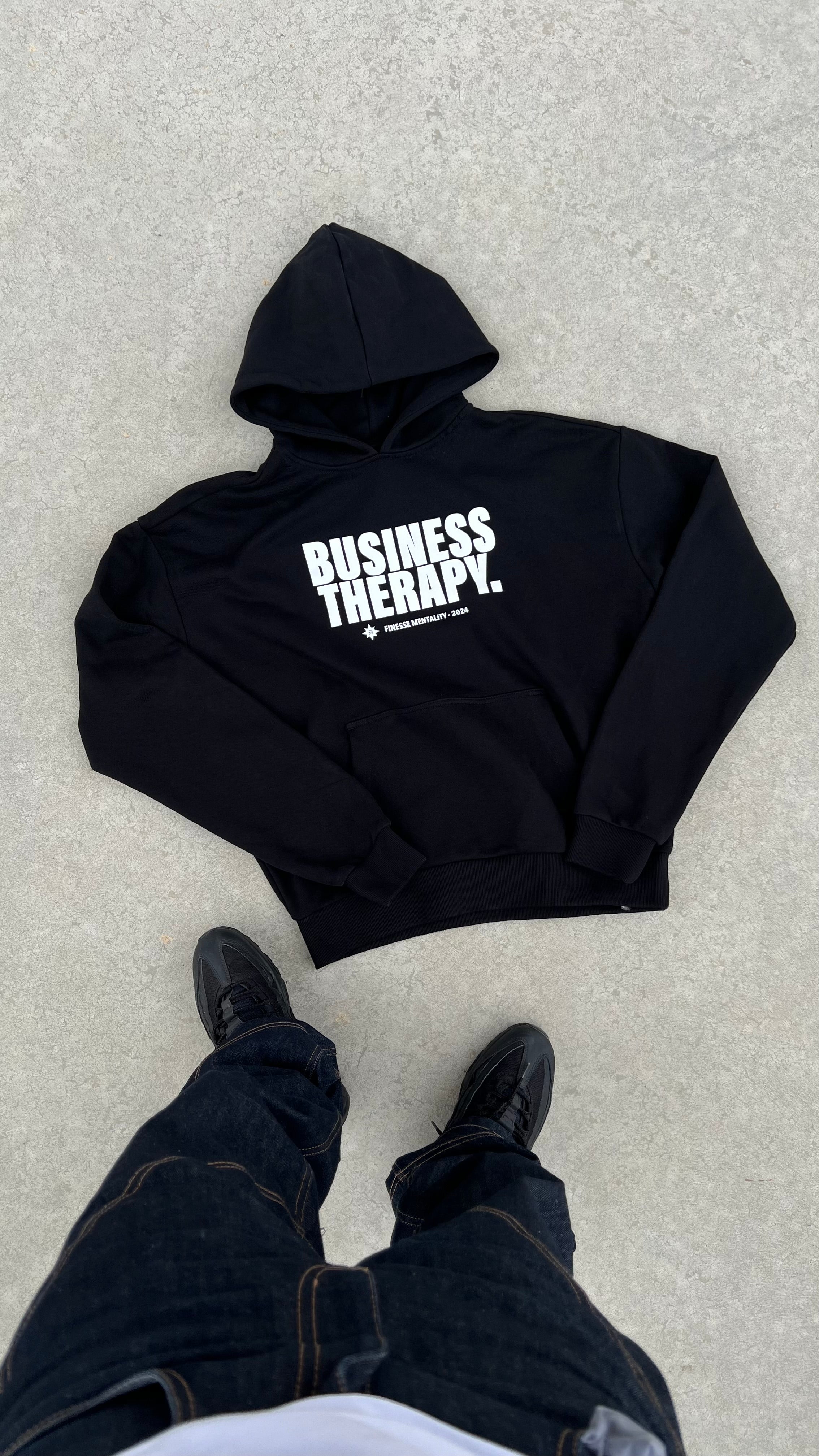 Hoodie Business Therapy Black de Finesse en packshot à plat, avec les pieds du photographe visibles.