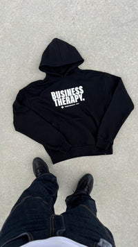 Hoodie Business Therapy Black de Finesse en packshot à plat, avec les pieds du photographe visibles.