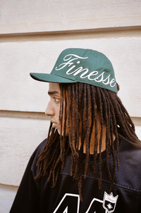 Homme portant une Finesse Sign Cap verte, regard à droite devant un mur.