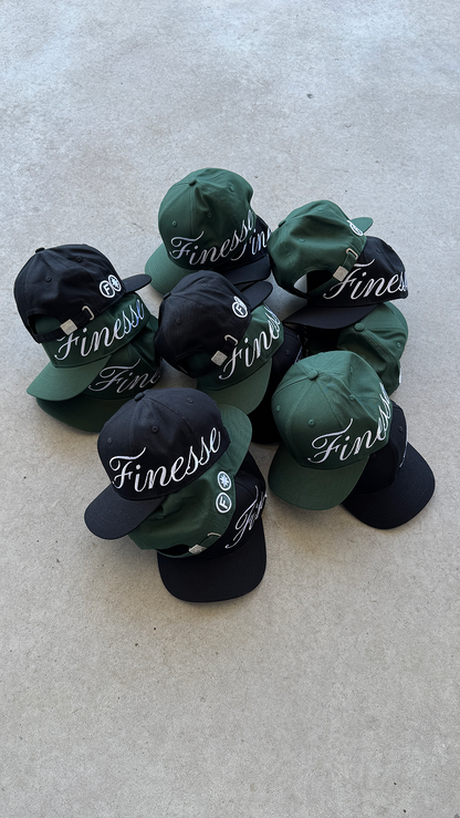 Plusieurs casquettes Finesse Sign Cap noires et vertes posées au sol en packshot.