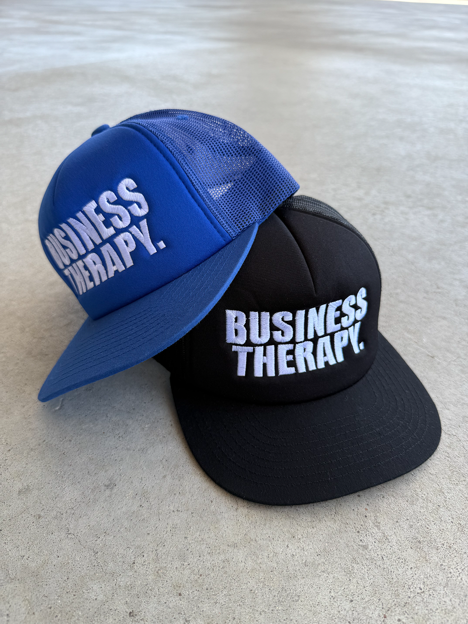 Deux casquettes Business Therapy Trucker, bleue et noire, présentées au sol.